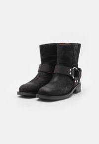 Botines de ante negro que cuentan con una amplia correa con herrajes plateados, superficie texturizada y un tacón bajo en bloque. Construcción simple con costuras.