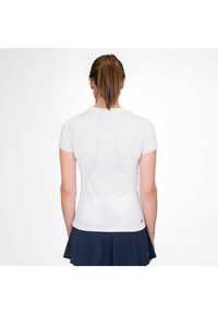Mujer con cola de caballo marrón, vistiendo una camiseta blanca ajustada de manga corta y una falda plisada azul marino, de pie de espaldas contra un fondo blanco.