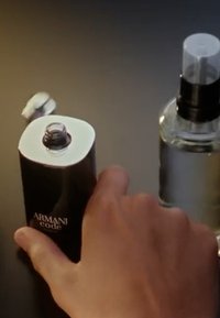 Hand reaching toward open Armani Code perfume bottle on left, next to a clear spray bottle with black pump on a dark surface.