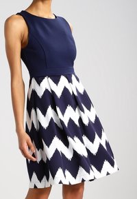 Mörkblå ärmlös klänning med en figursydd liv och en utsvängd kjol, med ett vitt zigzag-mönster. Materialet verkar slätt och lätt.