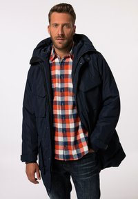 Marinblå vadderad jacka med huva, som har flera framfickor och en slät yta, bärs över en röd och blå rutig skjorta.