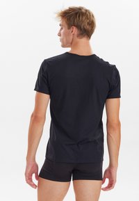 Sort kortærmet T-shirt i bomuld med rund halsudskæring, glat tekstur og tætsiddende design. Båret med sorte shorts, set fra bagenden.