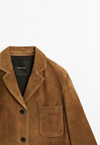Giacca in suede marrone con colletto a punta, chiusura frontale con bottoni e una tasca sul petto. Superficie texture con una finitura morbida.