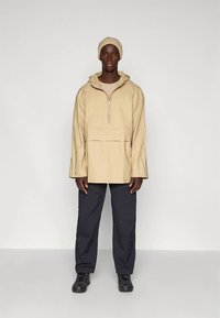 Beige pulloverjack met een halve ritssluiting, capuchon en wijde mouwen, gecombineerd met marineblauwe broek en zwarte schoenen, voor een gelaagde uitstraling.