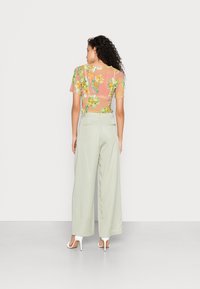 Túnica floral transparente em pêssego com flores verdes e amarelas; combinada com calças largas verde-claro e sapatos de salto altos brancos.