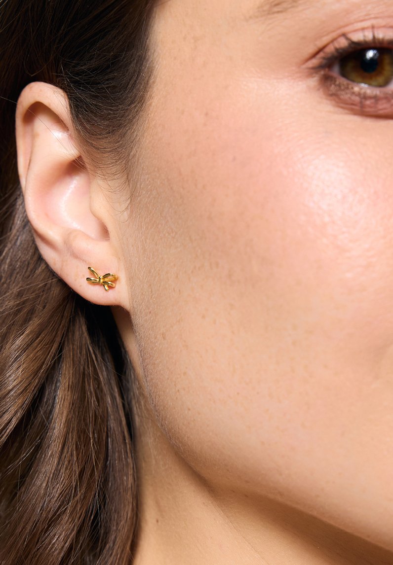 Złote kolczyki w kształcie motyla z teksturowanymi skrzydłami, charakteryzujące się delikatnym, misternym wzorem. Umiejscowione na małżowinie usznej modelki.