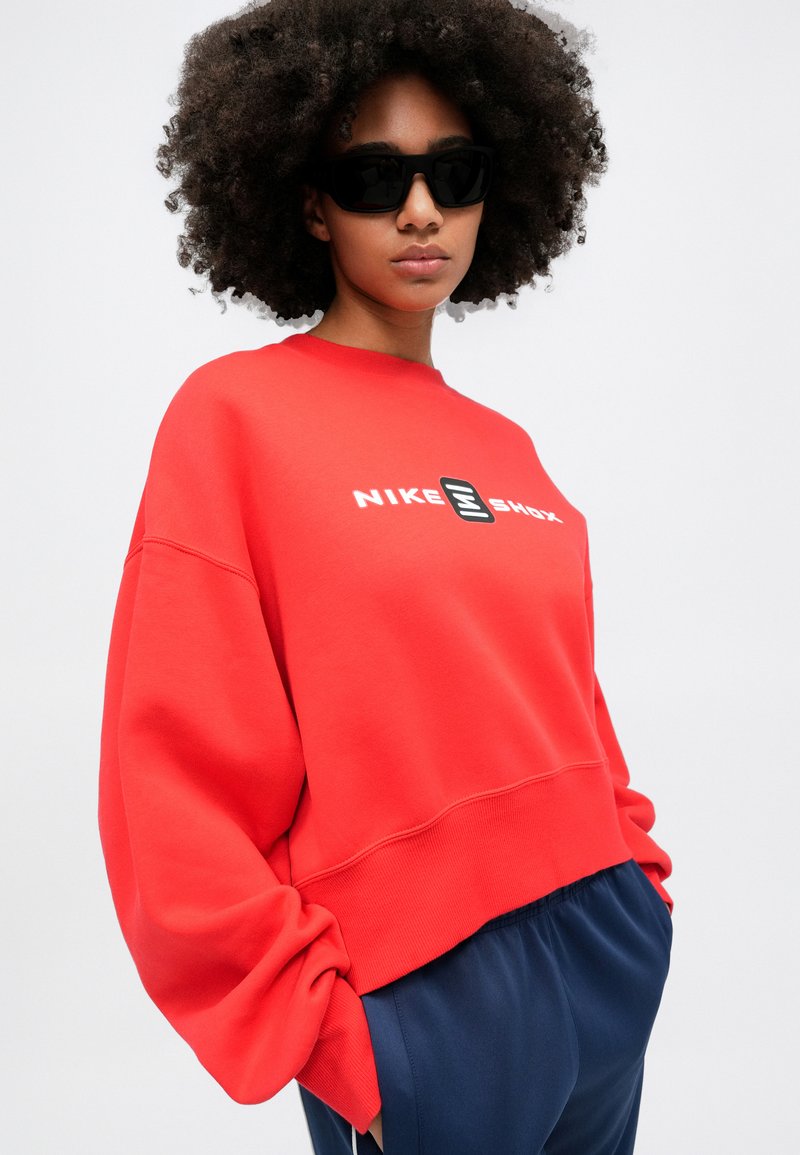 Felpa rossa Nike con colletto a girocollo, taglio oversize e logo bianco. Il modello indossa occhiali da sole neri, abbinati a pantaloni blu navy.