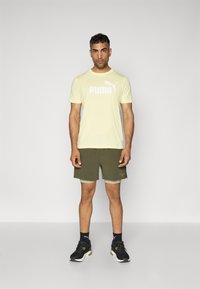 PUMA gul t-skjorte med korte ermer og hvit logo, kombinert med olivengrønne shorts og svarte treningssko. Enkelt design, uformell passform.