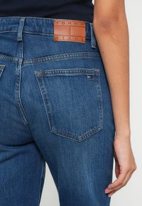 Tumed sinised teksapüksid, millel on pruuni nahaga vöökoha silt, millel on kiri "TOMMY JEANS". Omadusteks on kaks tõmbeauguga tagataskut õmblustega.