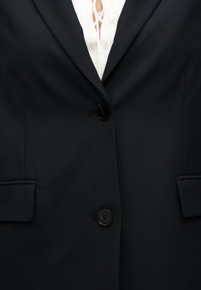 Μαύρο σακάκι ραμμένο καθ' όλη τη γραμμή με δύο εμπρόσθιες τσέπες, κλείσιμο με κουμπιά και ομαλή υφή υφάσματος. Κάτω από αυτό υπάρχει λευκό τοπ με δαντελένιο διακοσμητικό.