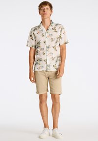 Camisa de manga corta, con botones, en tela ligera con un estampado floral en verde, naranja y beige. Combinada con pantalones cortos beige enrollados y zapatillas blancas.