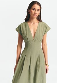 Olivgrön klänning med djup V-ringning och korta ärmar, med en åtsittande bodice och plisserad kjol, tillverkad av lätt tyg. Guldarmband som accent.