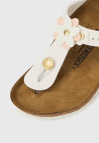 Bela sandal z rožnatimi in belimi cvetličnimi poudarki. Ima mehko podplat, nastavljiv trak in zlato gumbno podrobnost.