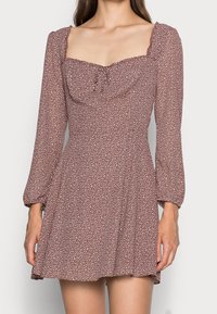 Mujer vestida con un vestido marrón de manga larga con lunares blancos y escote cuadrado, de pie contra un fondo liso.