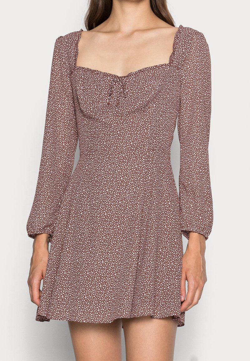 Mujer vestida con un vestido marrón de manga larga con lunares blancos y escote cuadrado, de pie contra un fondo liso.