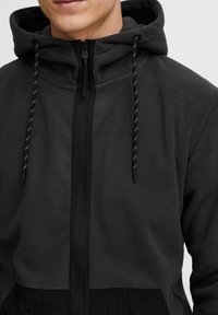 Fleece zip-jakke i mørkegrå med sorte detaljer. Har en hætte, justerbare snore og en frontlomme. Blød tekstur.