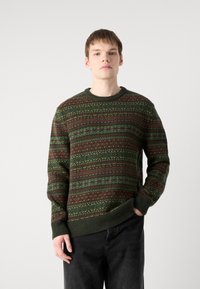 Knit sweater in dark green with multicolored horizontal patterns in red, yellow, and blue. Ribbed crew neckline and cuffs, paired with dark jeans.