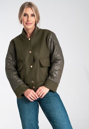 Olive green bomber jacket with quilted sleeves, two front pockets, and button closure; features contrasting textures and tailored design.