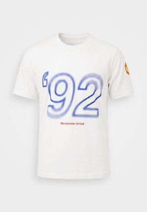 Biała koszulka Manchester United z dużym niebieskim napisem '92' z przodu oraz herbem klubu na prawym rękawie.