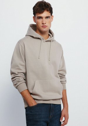 Haut à capuche en coton beige clair avec une poche kangourou, une capuche à cordon de serrage et des détails de poignets côtelés, porté sur un jean en denim foncé.