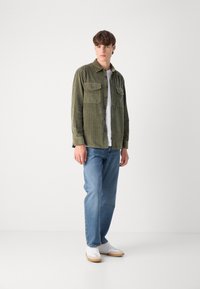 Groene corduroy shirt met twee borstzakken, gecombineerd met blauwe spijkerbroeken en witte sneakers met beige zolen. Model staat tegen een effen achtergrond.