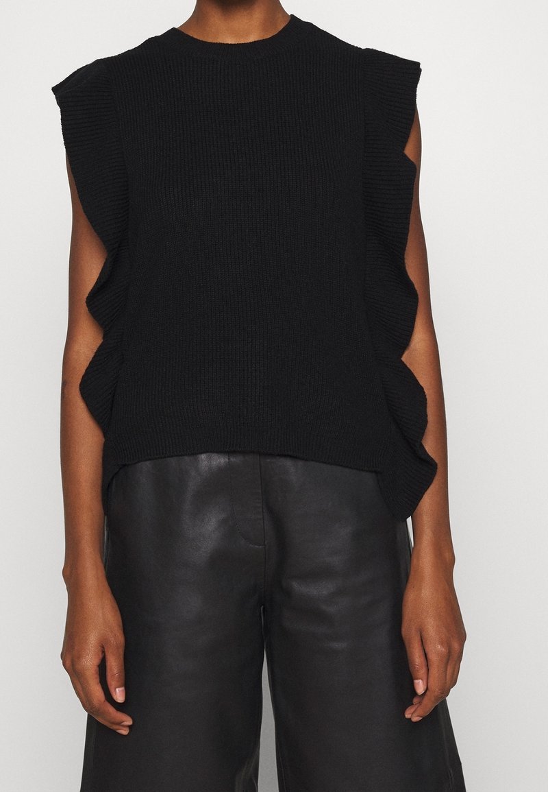 Haut en maille noir sans manches avec côtés volantés porté avec un pantalon en cuir noir, montré du cou jusqu'à la mi-cuisse.