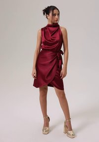 Γυναίκα φορώντας ένα φόρεμα από βουργουνδερίνι σατέν χωρίς μανίκια και χρυσές πλατφόρμες, στέκεται με χέρια χαλαρά, κοιτάζοντας προς το πλάι.