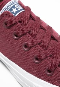 Zbliżenie na bordowy materiałowy sneaker Converse z dopasowanymi sznurowadłami i białą gumową podeszwą, z widocznym logo gwiazdy na języku.