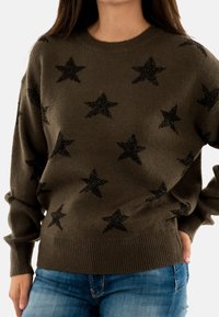 Donna che indossa un maglione lavorato a maglia marrone con stelle nere abbinato a jeans blu, con la mano appoggiata sull'anca, su sfondo bianco.