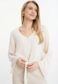Blusa ligera de color crema con escote en V, mangas largas de campana y detalle de lazo en la parte delantera; presenta cierres de botón a lo largo del escote.