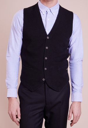 Ein Mann trägt eine schwarze, geknöpfte Weste über einem hellblauen Langarmhemd mit passenden schwarzen Hosen und steht vor einem einfarbigen Hintergrund.