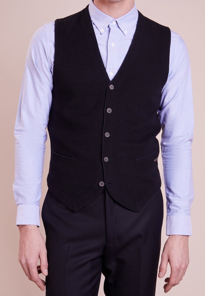 Homme portant un gilet noir boutonné par-dessus une chemise bleu clair à manches longues avec un pantalon noir assorti, debout devant un fond uni.
