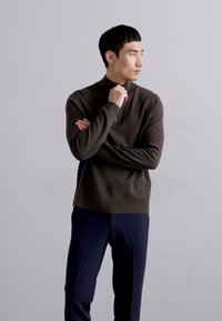Uomo vestito con una camicia a maniche lunghe marrone scuro e pantaloni blu navy, in piedi con le braccia incrociate e che guarda verso destra su uno sfondo semplice.