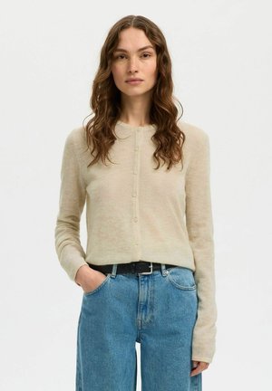 Femme aux longs cheveux ondulés portant un cardigan beige boutonné rentré dans un jean bleu taille haute avec une ceinture noire, une main dans la poche.