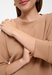 Khakifarbenes Sweatshirt mit Rundhalsausschnitt und Dreiviertelärmeln. Glatte Textur; zeigt sichtbare Nahtdetails. Goldene Kreolenohrringe sind sichtbar.