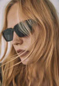 Musta suorakulmion muotoiset aurinkolasit tummilla linsseillä, joissa on kiiltävä pinta ja hienovaraiset kultaiset yksityiskohdat sangan kohdalla. Hiukset valuvat kehysten ympärillä.