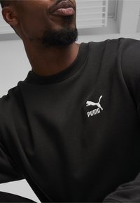 Felpa nera con collo a girocollo a costine e logo Puma ricamato di colore bianco sul lato sinistro superiore. Texture morbida e design casual.