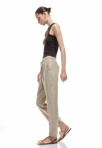Top de tirantes marrón con relieve, pantalones beige holgados con bolsillos y sandalias marrones con tiras. El modelo está de perfil con una postura relajada.