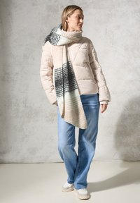 Beige Daunenjacke mit steppdesign, kombiniert mit einem langen Schal im Karomuster in Pink, Schwarz und Creme. Hellblaue Jeans und graue Sneakers.