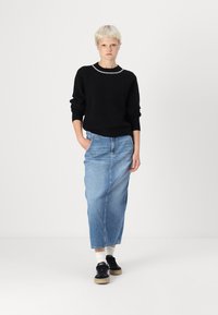 Črna rebrasta pletena srajca z belim robom, v kombinaciji z svetlo modrim denim krilom in črnimi supergami s svetlo rjavim podplatom. Model stoji z rokami v žepih.