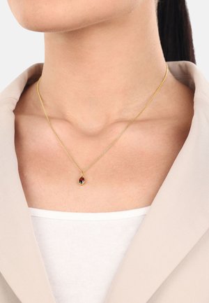 Goldkette mit einem ovalen roten Stein, feiner Kette, über einem hellen Oberteil getragen, teilweise sichtbar unter einer beigen Jacke.