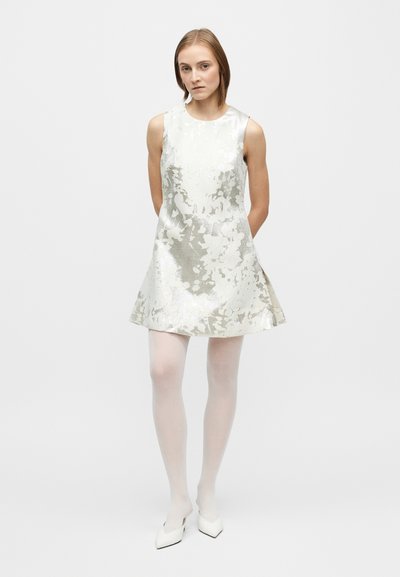 Rochie argintie fără mâneci, cu imprimeu floral și fustă evazată, confecționată din material reflectorizant, asortată cu colanți albi și pantofi cu vârf ascuțit.