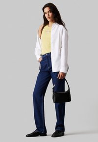 Camisa blanca de botones sobre una camiseta sin mangas amarilla clara, jeans azules y zapatos planos negros. Sostiene un pequeño bolso negro en una mano.
