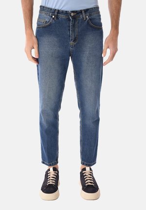 Uomo che indossa jeans slim fit blu medio e sneaker nere con lacci bianchi, in piedi davanti a uno sfondo bianco.