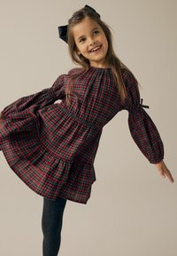 Rød og grønternet kjole med puffedeærmer, smocket liv og tiered skirt. Båret med grå teksturerede strømpebukser. Sort sløjfe i håret.