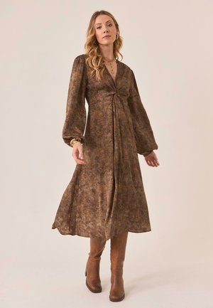 Mujer con un vestido largo marrón con estampado, mangas largas abullonadas y botas de cuero marrón hasta la rodilla, de pie sobre un fondo liso.