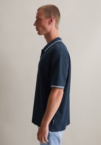 Ciemnoniebieska koszulka polo z teksturowanej tkaniny, z kołnierzykiem w białe paski i krótkimi rękawami. Luźny krój, z bocznymi rozcięciami.