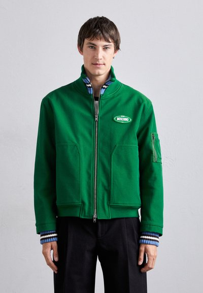 Zielona kurtka z zippem, z dwiema przednimi kieszeniami, paskowanym niebieskim kołnierzem oraz logo Moschino na lewej piersi. Posiada kieszeń na rękawie zapinaną na zamek.