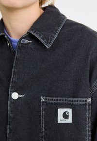 Črna denim jakna s koničastim ovratnikom, belim zapiranjem na gumb in žepom s krpico, na kateri je logotip. Očitne podrobnosti kontrastnega šivanja.