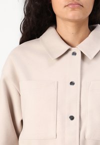 Camicia a bottoni beige chiaro realizzata in tessuto testurizzato, dotata di collo, due tasche frontali e chiusure a pressione.